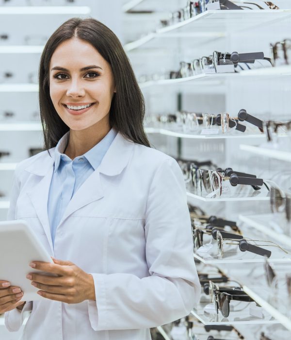 beautiful smiling optician using digital tablet near shelves with eyeglasses in ophthalmic shop